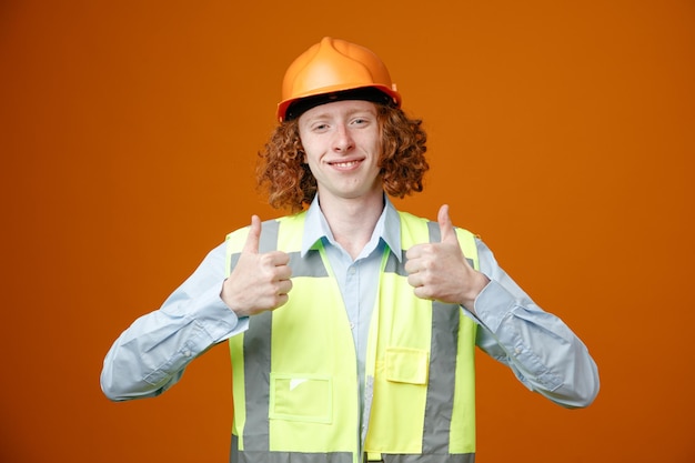 Joven constructor en uniforme de construcción y casco de seguridad mirando a la cámara feliz y positivo sonriendo alegremente mostrando los pulgares hacia arriba de pie sobre fondo naranja