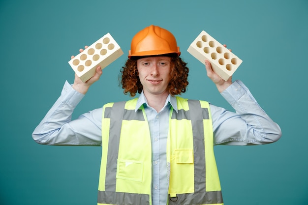 Joven constructor en uniforme de construcción y casco de seguridad sosteniendo dos ladrillos mirando a la cámara con una sonrisa en la cara de pie sobre fondo azul.