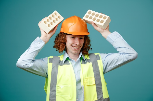 Joven constructor con uniforme de construcción y casco de seguridad sosteniendo dos ladrillos sobre su cabeza sonriendo alegremente mirando a la cámara de pie sobre fondo azul