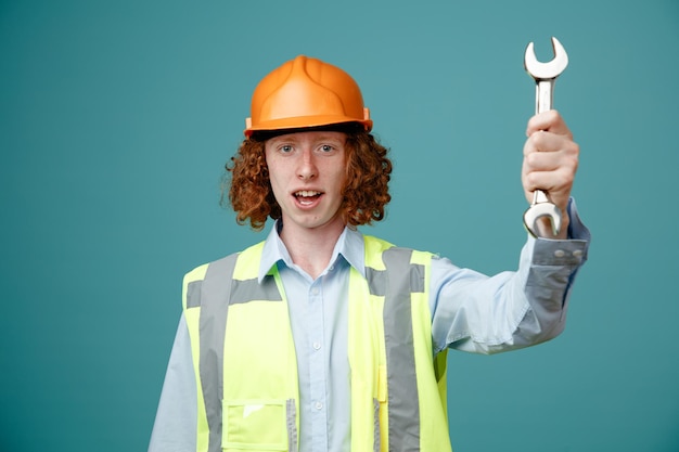 Joven constructor en uniforme de construcción y casco de seguridad sosteniendo una llave mirando a la cámara emocionado de pie sobre fondo azul.