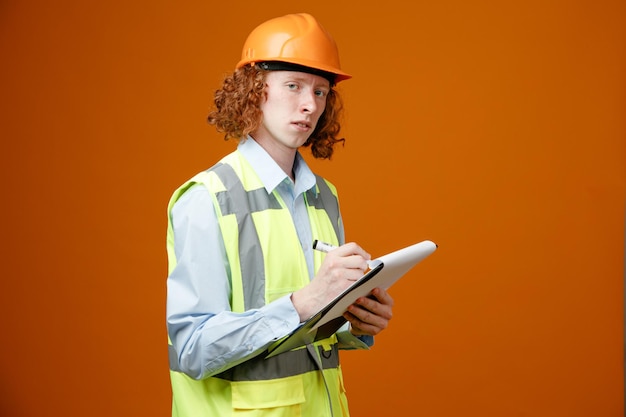 Joven constructor en uniforme de construcción y casco de seguridad sosteniendo portapapeles y marcador haciendo notas mirando confiado de pie sobre fondo naranja