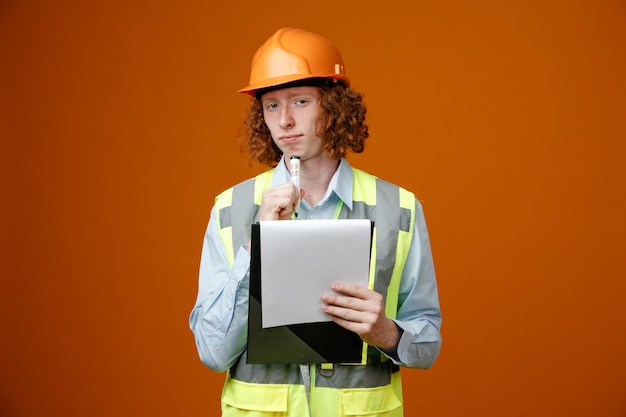 Foto joven constructor en uniforme de construcción y casco de seguridad sosteniendo portapapeles y marcador mirando a la cámara con cara seria de pie sobre fondo naranja