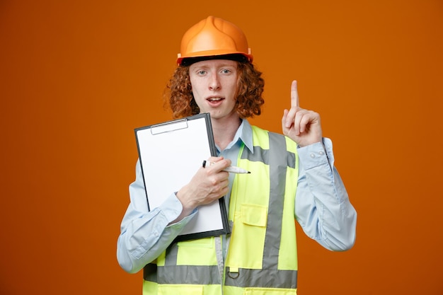 Joven constructor en uniforme de construcción y casco de seguridad sosteniendo portapapeles y marcador mirando a la cámara feliz y positivo mostrando el dedo índice con una nueva idea de pie sobre fondo naranja