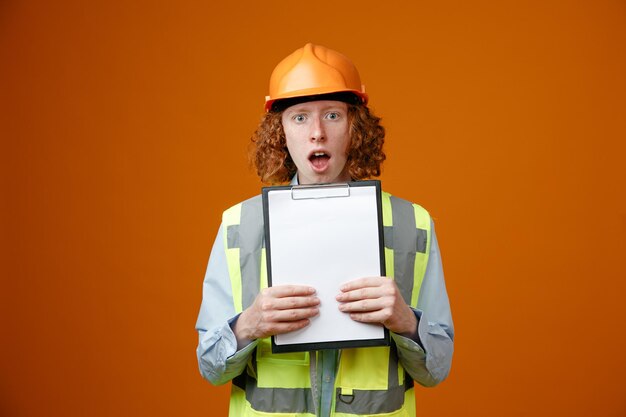 Joven constructor en uniforme de construcción y casco de seguridad sosteniendo portapapeles con páginas en blanco mirando a la cámara sorprendido y asombrado de pie sobre fondo naranja