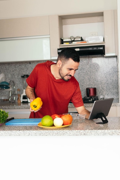 Joven consultando una receta en línea con una tableta mientras prepara una comida en la cocina de su casa