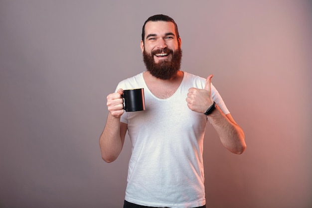 El joven contenido sostiene una taza negra mientras muestra el pulgar hacia arriba para dar me gusta