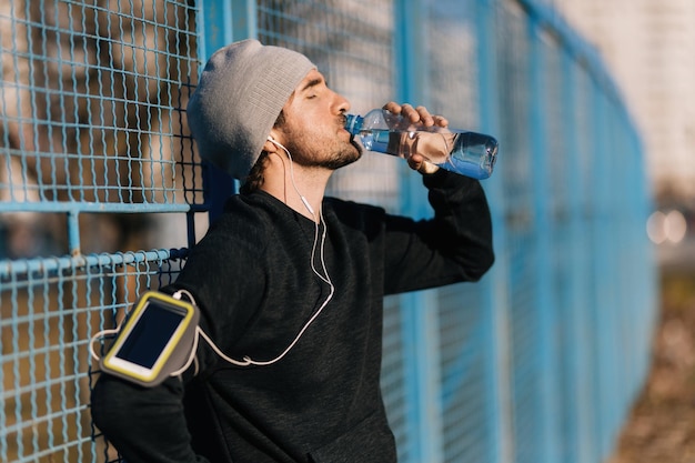 Joven corredor bebiendo agua de una botella mientras se apoya en una valla al aire libre