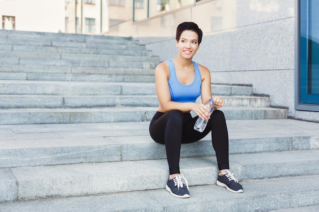 La joven corredora está tomando un descanso, bebiendo agua mientras trota en la ciudad, sentada en las escaleras y sonriendo, copiando espacio