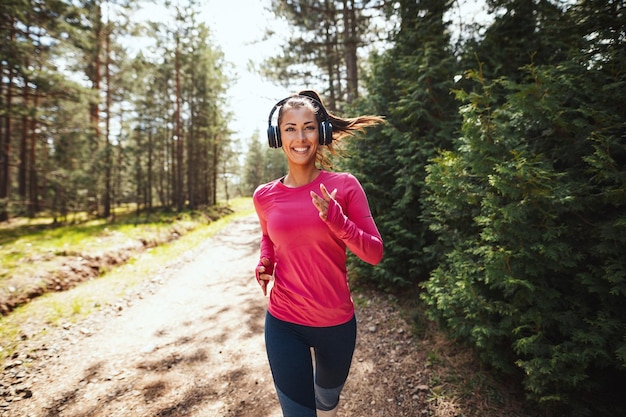 Joven corredora feliz escuchando música mientras trota por un sendero soleado en el bosque.