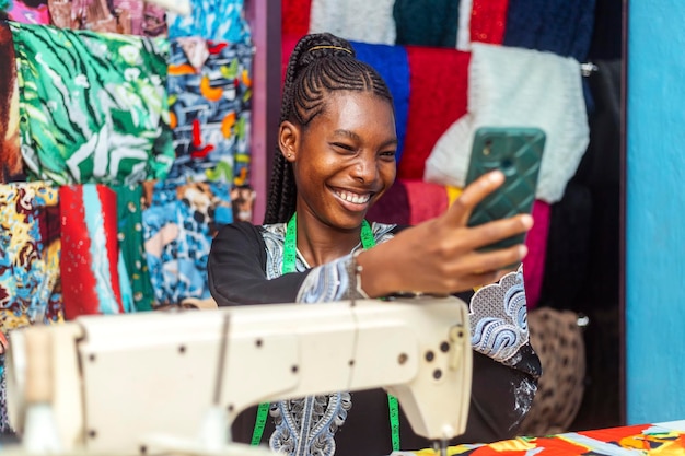 Foto joven costurera africana feliz sonríe usando el teléfono móvil mientras está aislada sobre el fondo de la ropa