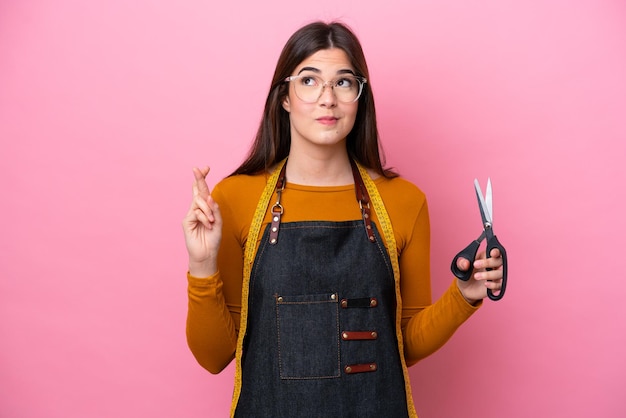 Joven costurera brasileña aislada de fondo rosa con los dedos cruzados y deseando lo mejor