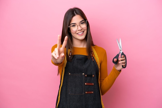 Joven costurera brasileña aislada de fondo rosa sonriendo y mostrando el signo de la victoria