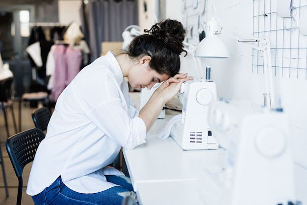 Joven costurera cansada o enferma sentada junto al escritorio y manteniendo la cabeza en la máquina de coser eléctrica en el taller