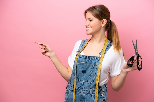 Joven costurera caucásica aislada de fondo rosa señalando con el dedo hacia un lado y presentando un producto