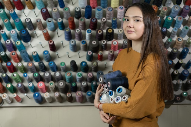 Una joven costurera en un taller se encuentra cerca de un puesto con hilos de diferentes colores. Tiene varias madejas en sus manos. La elección del material de sutura para coser una camisa. Copiar espacio.