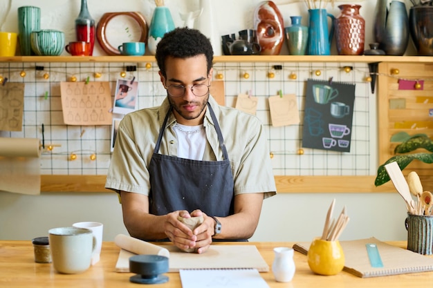 Joven creativo experto en hacer loza sentado junto a la mesa en el taller