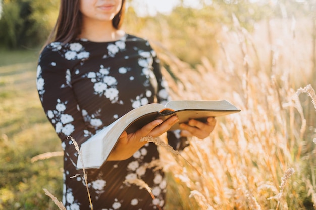 Joven cristiana religiosa sosteniendo y leyendo su biblia, afuera en el campo al atardecer