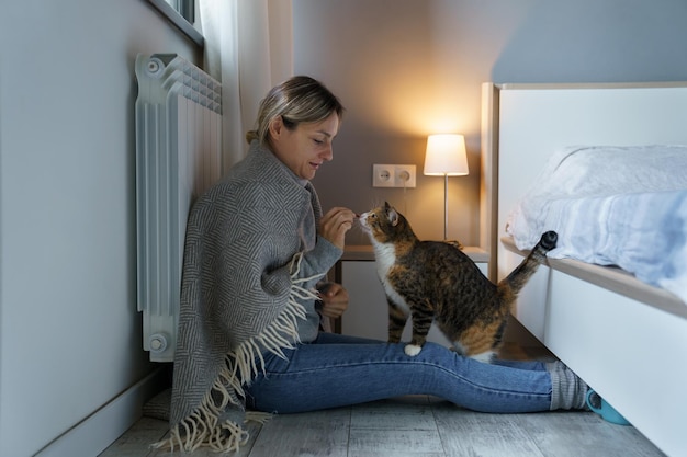 Una joven cubierta con una manta caliente juega con un gato alimentando a un animal con comida sentada en el suelo