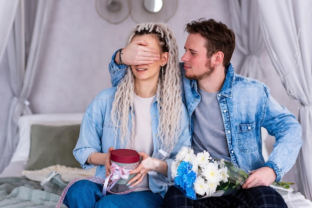 El joven cubre los ojos de la chica con la mano y le da flores La chica está sentada en la cama con un regalo en las manos Joven pareja alegre con ropa informal