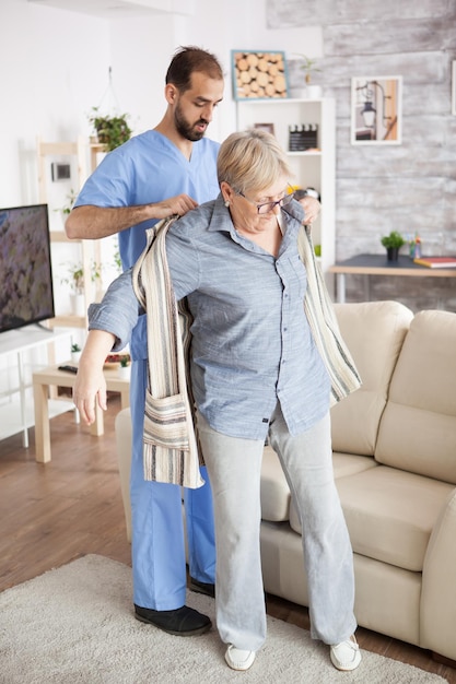 Un joven cuidador masculino en un hogar de ancianos ayudando a una anciana a vestirse.