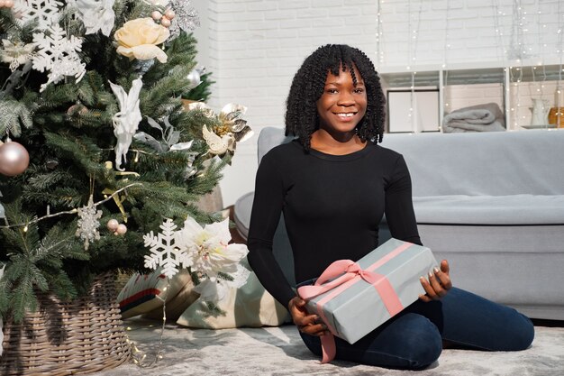 Joven dama afroamericana se sienta cerca de un árbol de Navidad y tiene un regalo