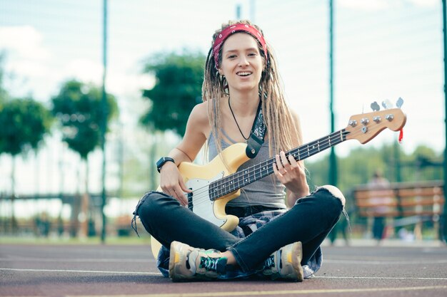 Joven dama de pelo largo con rastas sentada con las piernas cruzadas y sonriendo mientras toca la guitarra