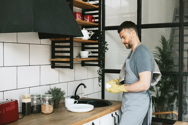 un joven con delantal y guantes hace tareas domésticas lava platos con una toalla en el hombro hombres que hacen tareas domésticas ayudan en una cocina elegante en un apartamento moderno