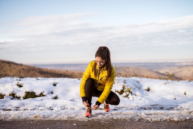 Joven delgada chica atlética atar cordones de los zapatos en ropa deportiva de invierno en camino de invierno cubierto de nieve con auriculares en la mañana soleada.