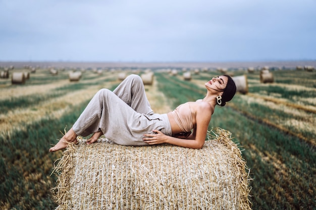 Joven delgada morena con maquillaje brillante en top beige con estómago desnudo y pantalones grises, acostado en el heno, posando, sesión de fotos en el campo, cielo azul.