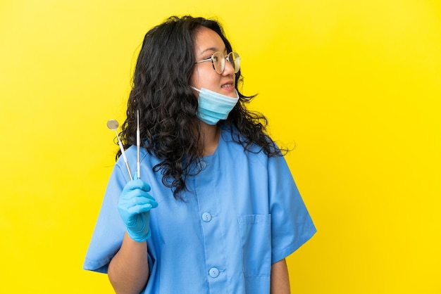 Joven dentista asiático sosteniendo herramientas sobre antecedentes aislados mirando hacia el lado y sonriendo