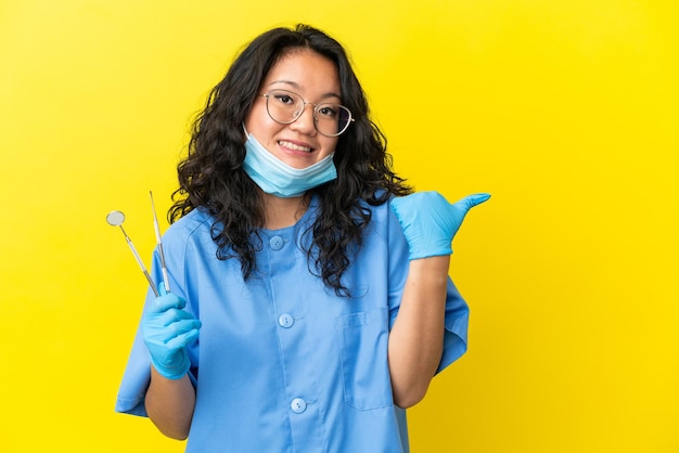 Joven dentista asiático sosteniendo herramientas sobre fondo aislado apuntando hacia el lado para presentar un producto