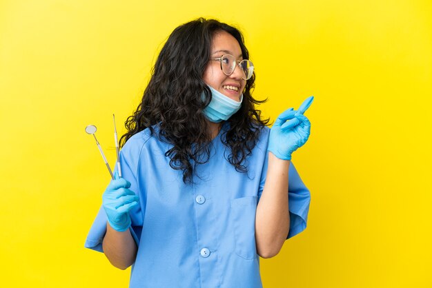 Joven dentista asiático sosteniendo herramientas sobre fondo aislado con la intención de darse cuenta de la solución mientras levanta un dedo