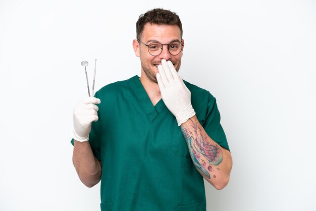 Joven dentista caucásico aislado de fondo blanco feliz y sonriente cubriendo la boca con la mano