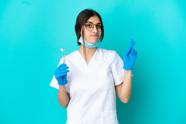 Joven dentista caucásico mujer sosteniendo herramientas aisladas sobre fondo azul con los dedos cruzando y deseando lo mejor