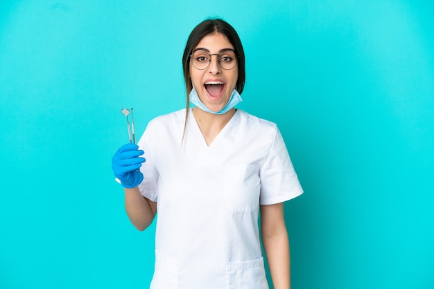 Joven dentista caucásico mujer sosteniendo herramientas aisladas sobre fondo azul con expresión facial sorpresa