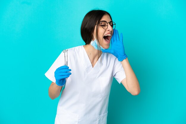 Joven dentista caucásico mujer sosteniendo herramientas aisladas sobre fondo azul gritando con la boca abierta hacia el lado