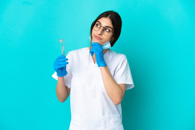 Joven dentista caucásico mujer sosteniendo herramientas aisladas sobre fondo azul y mirando hacia arriba