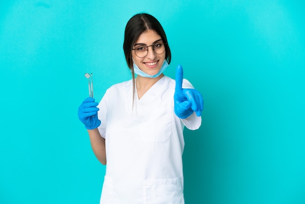 Joven dentista caucásico mujer sosteniendo herramientas aisladas sobre fondo azul mostrando y levantando un dedo