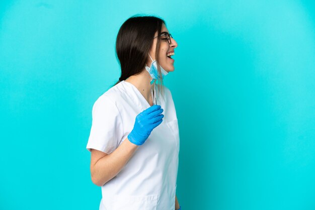 Joven dentista caucásico mujer sosteniendo herramientas aisladas sobre fondo azul riendo en posición lateral