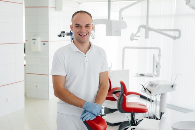 Un joven dentista se para cerca de un sillón dental rojo y sonríe en la odontología blanca moderna Tratamiento y prevención de caries desde la juventud Odontología moderna y prótesis