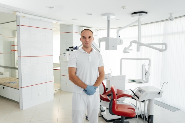 Un joven dentista se para cerca de un sillón dental rojo y sonríe en la odontología blanca moderna Tratamiento y prevención de caries desde la juventud Odontología moderna y prótesis