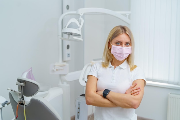 Joven dentista se encuentra con las manos cruzadas en la clínica de odontología moderna. Doctor con mascarilla.