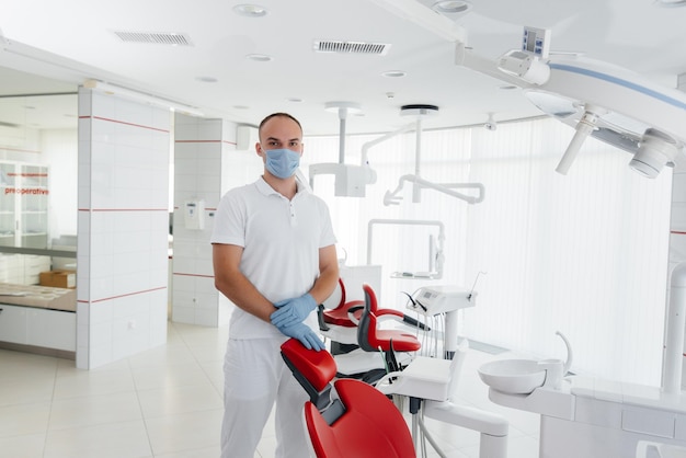 Un joven dentista con una máscara se para cerca de una silla dental roja y sonríe en la odontología blanca moderna Odontología moderna y prótesis