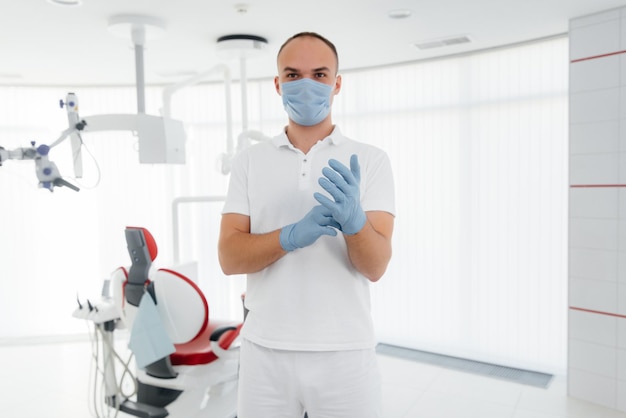 Un joven dentista con una máscara se para cerca de una silla dental roja y sonríe en la odontología blanca moderna Odontología moderna y prótesis