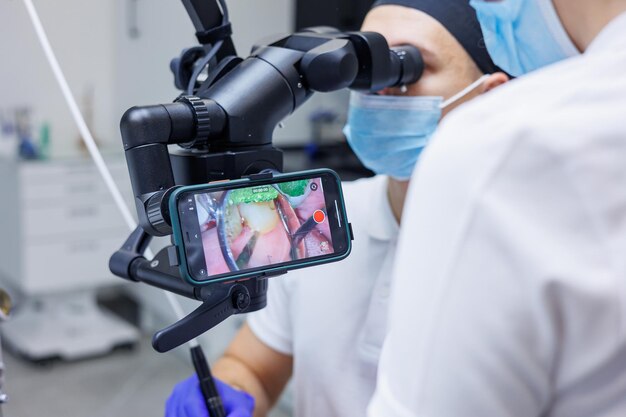 Un joven dentista mira los dientes del paciente con un microscopio dental y sostiene instrumentos dentales cerca de la boca Un asistente ayuda al médico Visten uniformes blancos con máscaras