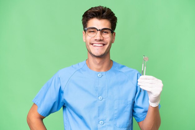 Joven dentista sosteniendo herramientas sobre un fondo aislado posando con los brazos en la cadera y sonriendo