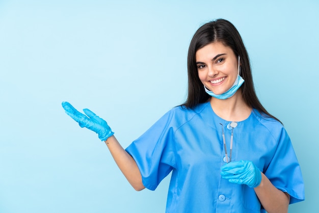 Joven dentista sosteniendo herramientas sobre pared azul aislada extendiendo las manos hacia un lado para invitar a venir