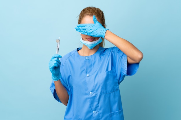 Joven dentista sosteniendo herramientas sobre pared azul aislada que cubre los ojos con las manos