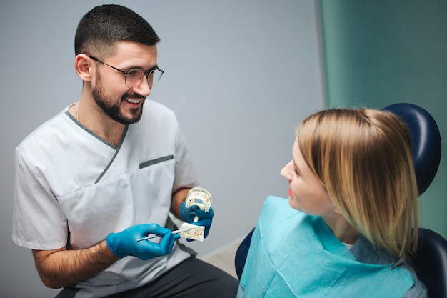 Joven dentista sosteniendo la mandíbula artificial con dientes y mostrándola a un paciente