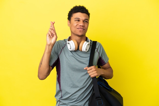 Joven deporte afroamericano con bolsa de deporte aislado sobre fondo amarillo con los dedos cruzando y deseando lo mejor